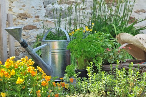 The image depicts a small garden corner with a variety of plants and gardening tools. In the foreground, there are bright yellow and orange flowering plants, possibly marigolds, adding vibrant colour to the space. Behind them, there is a lush green herb or leafy plant in a black container, with its fronds extending upward. To the right, a watering can made of metal is positioned slightly to the side, suggesting ongoing garden maintenance. Adjacent to the watering can, there is a metal scoop or garden trowel placed on the ground for planting or watering tasks. The background features a weathered stone wall, adding a rustic, natural backdrop to the scene. Tall grass or reeds grow along the brick wall, contributing to the lush, outdoor environment. A straw or woven garden hat rests on a surface near the plants, indicating active gardening activity. The overall setting appears to be a well-maintained, landscaped garden space typical of outdoor garden care in Anerley, with natural light suggesting a bright, partly sunny day, ideal for gardening activities. This detailed scene aligns with the services offered by Lawn Mowing Anerley, specializing in gardening and outdoor maintenance.