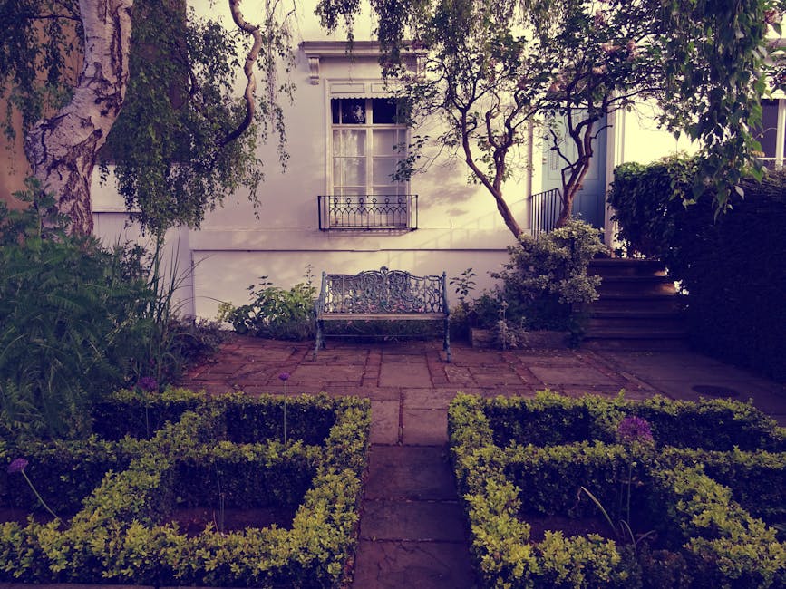 A neatly maintained garden in front of a white Victorian terrace house, featuring a paved pathway leading to an ornate metal garden bench centered against the house wall. The garden is bordered by low